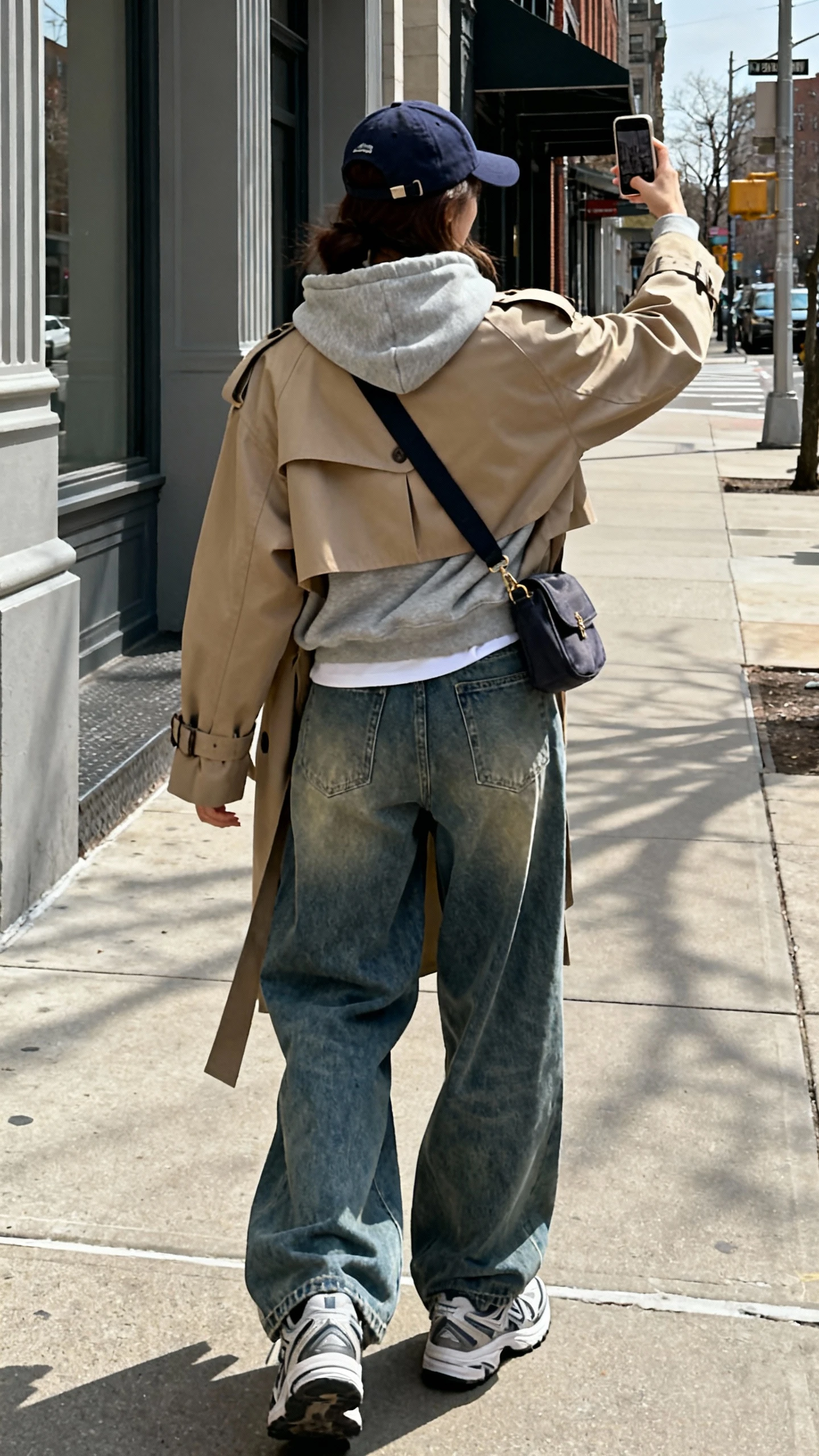 iPhone back-view street selfie of a woman in baggy jeans, a hoodie layered under a trench coat, sporty sneakers, baseball cap and crossbody bag, face not visible, natural daylight on city sidewalk, casual iPhone quality