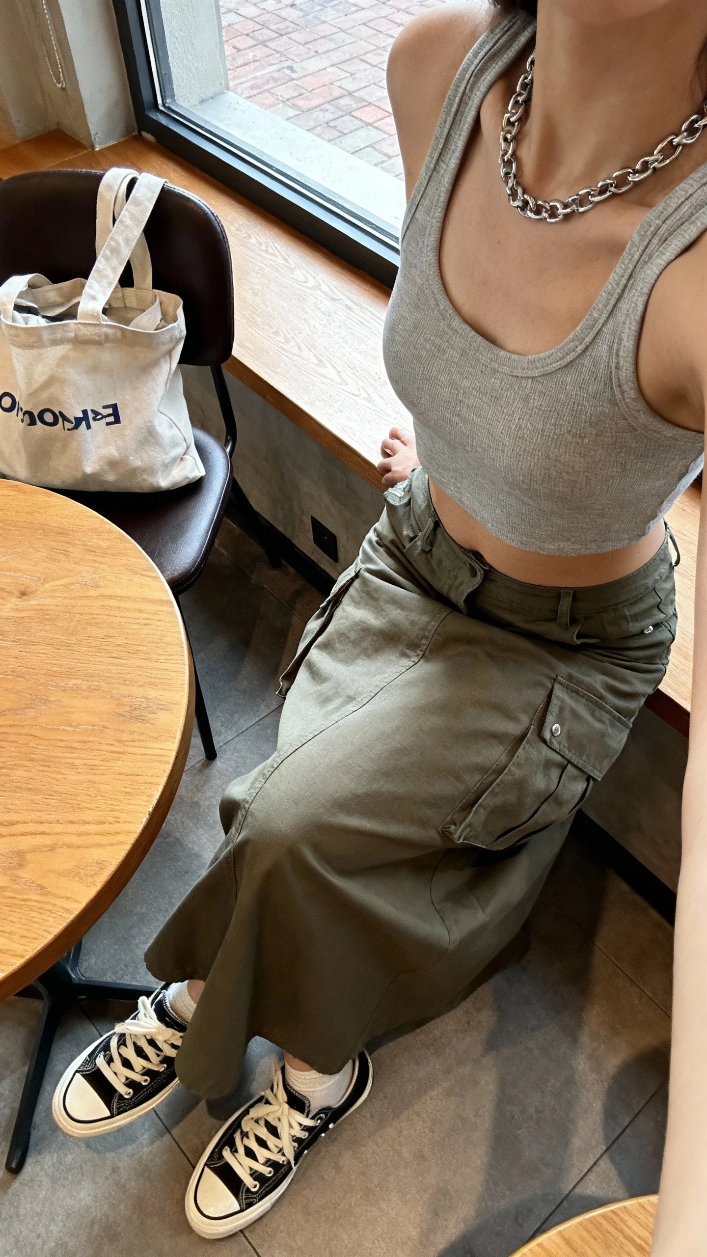 iPhone cafe table selfie cropped at shoulders showing a woman in a cropped tank with a cargo maxi skirt, low-profile skate sneakers, silver chain necklace, canvas tote on chair, face not visible, cozy coffee shop window seat, soft afternoon light, natural iPhone photo quality.