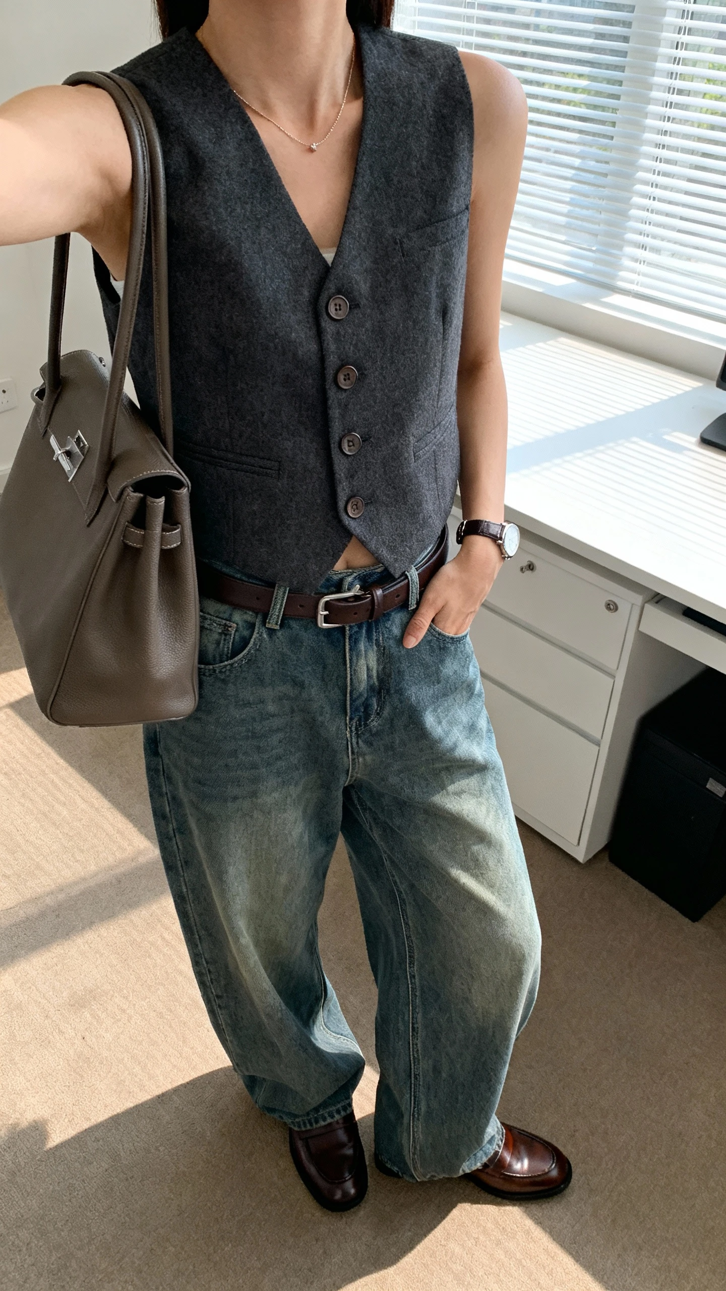 iPhone desk-side selfie cropped at neck showing a woman wearing baggy jeans, a buttoned waistcoat-style vest, polished loafers, structured tote, belt and delicate watch, face not visible, bright office window light, natural iPhone photo quality