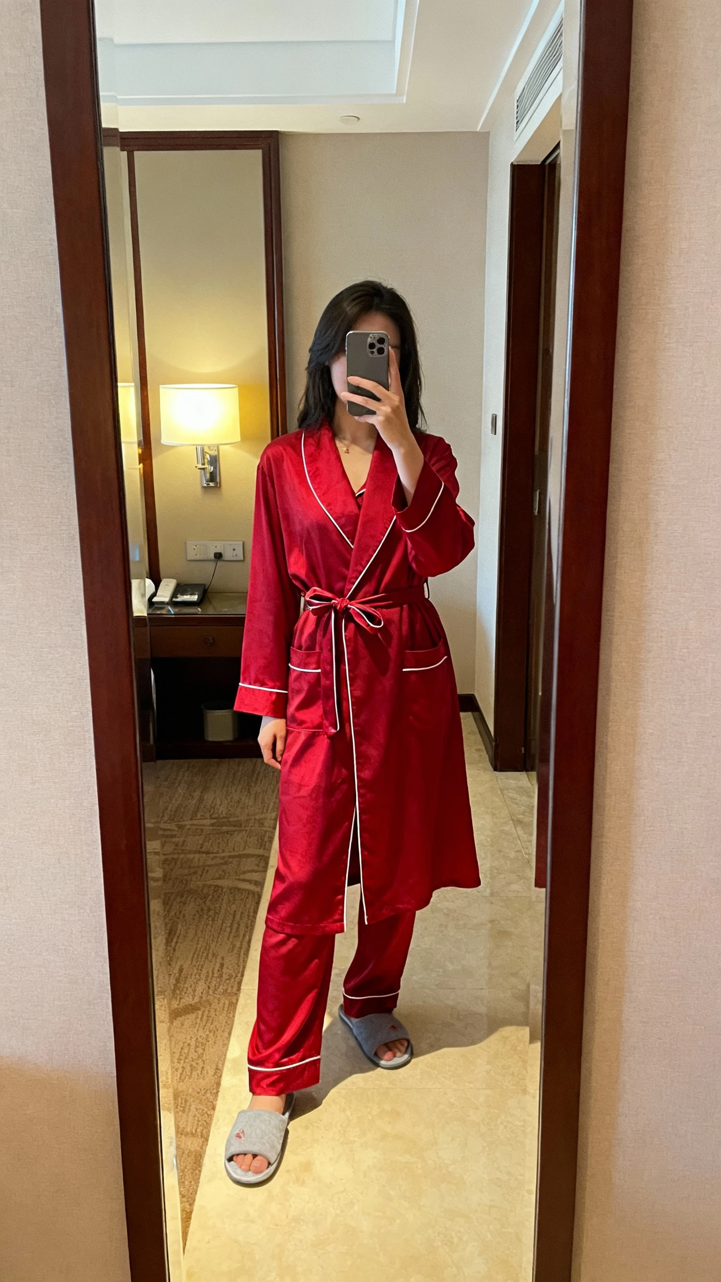 iPhone mirror selfie of a woman in a classic red pajama set with contrast piping, tied belted robe over matching top and pants, slippers, face not visible, hotel room full-length mirror, warm indoor lamp light, natural iPhone photo.