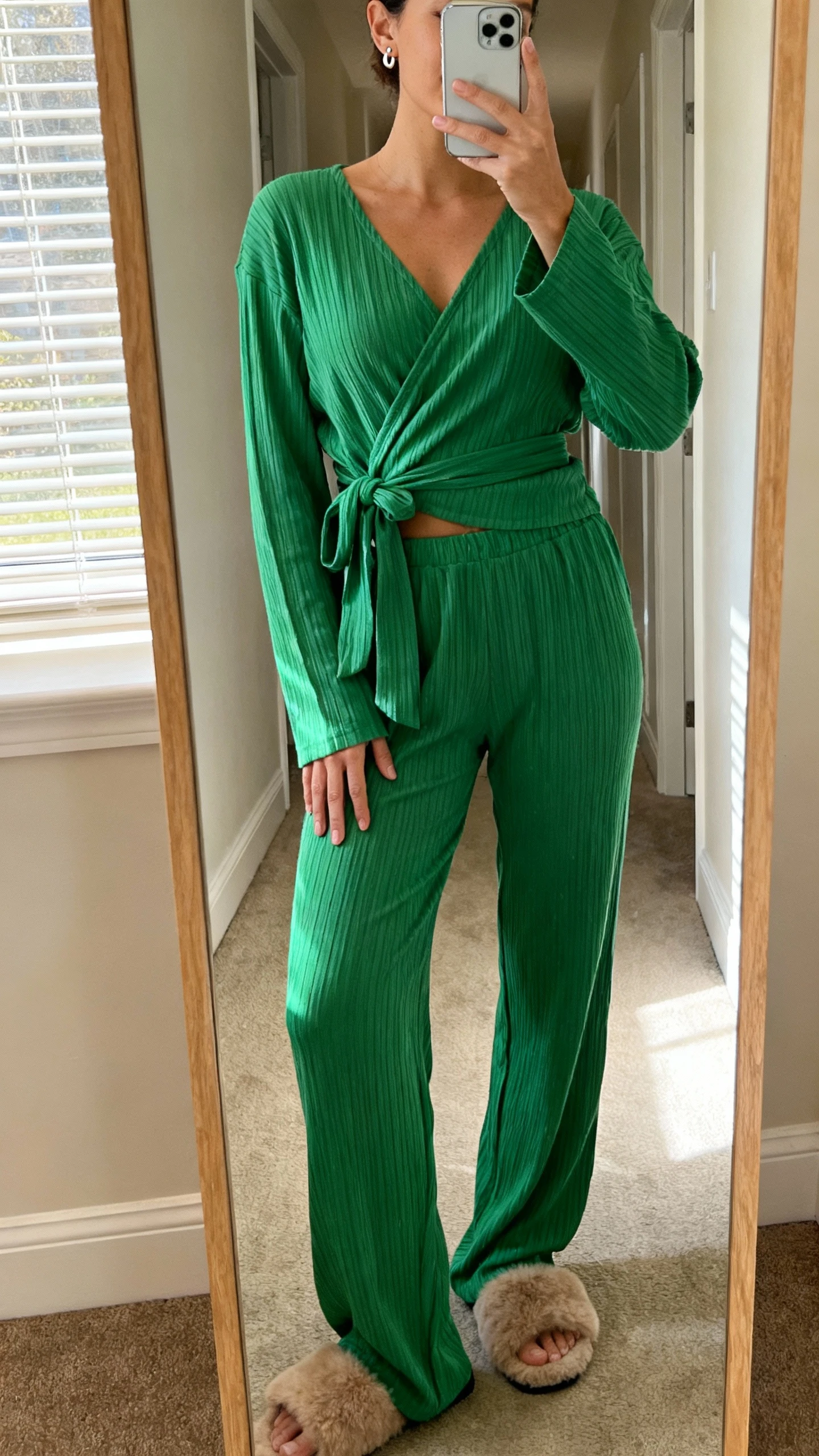 iPhone mirror selfie of a woman in an evergreen ribbed lounge set featuring a wrap top with tie and matching ribbed pants, simple stud earrings and fuzzy slippers, face not visible, hallway full-length mirror, morning window light, casual iPhone photo.