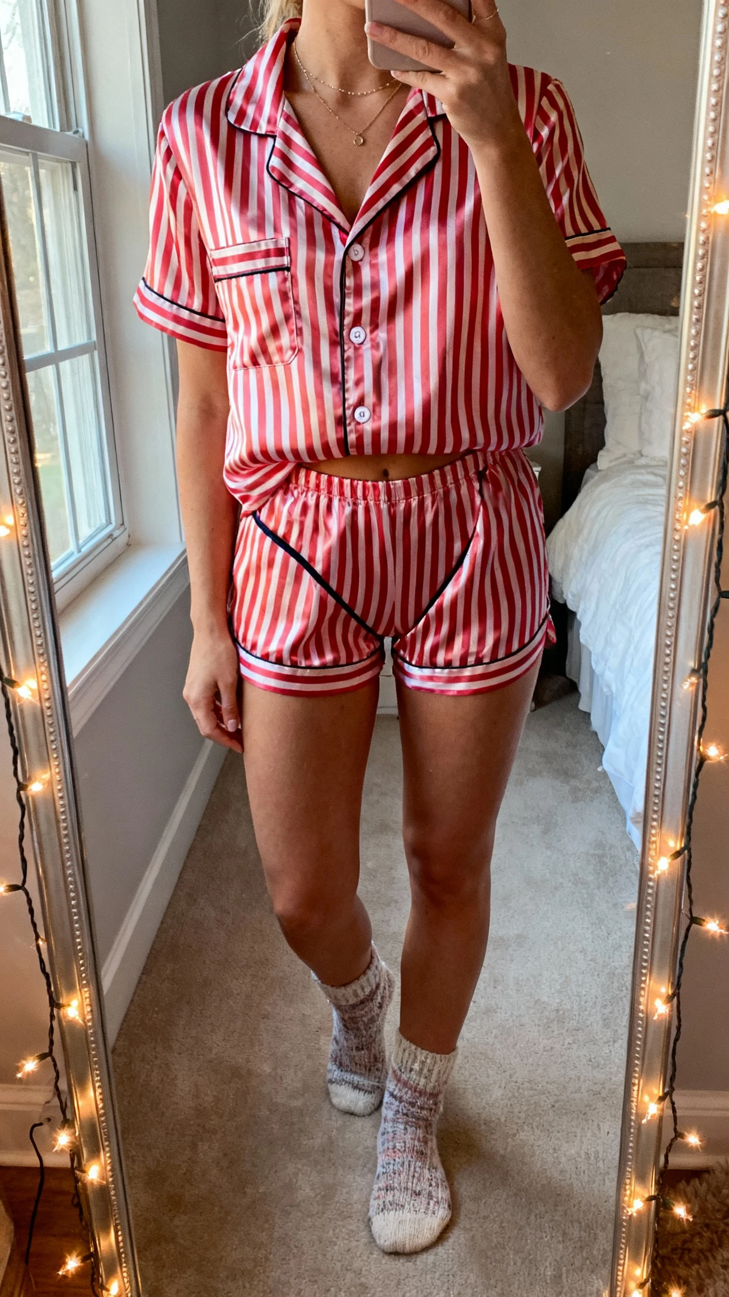 iPhone mirror selfie of a woman wearing a candy cane stripe satin pajama set with a button-up top and cheeky shorts, minimal jewelry and cozy socks, face not visible, cropped at neck, bedroom mirror with twinkle lights, soft window light, natural iPhone photo quality.