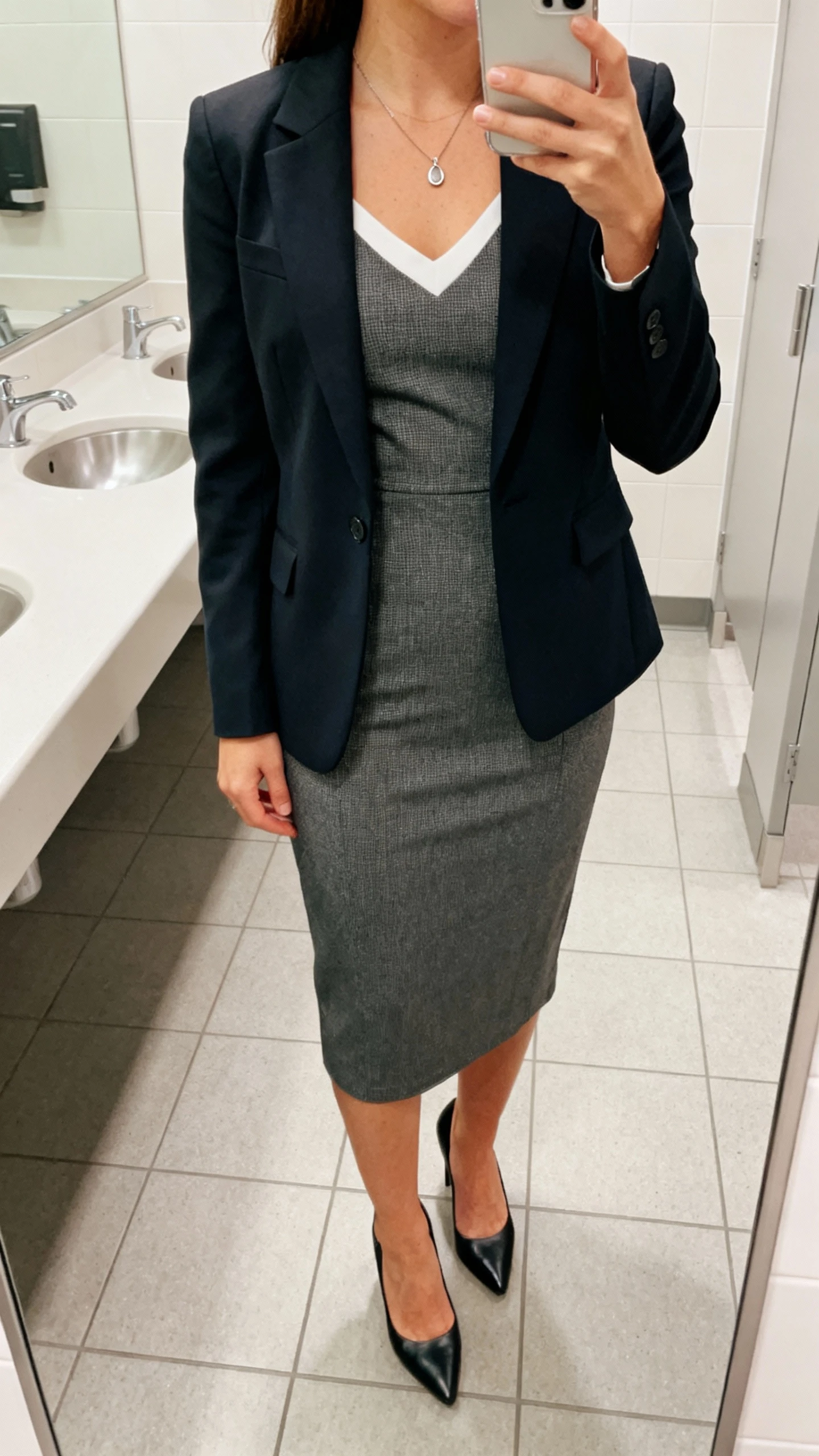 iPhone mirror selfie of a woman wearing a midi sheath dress layered with a tailored blazer, simple pendant necklace and classic pumps, face not visible, restroom mirror at work, bright indoor lighting, natural iPhone photo quality.
