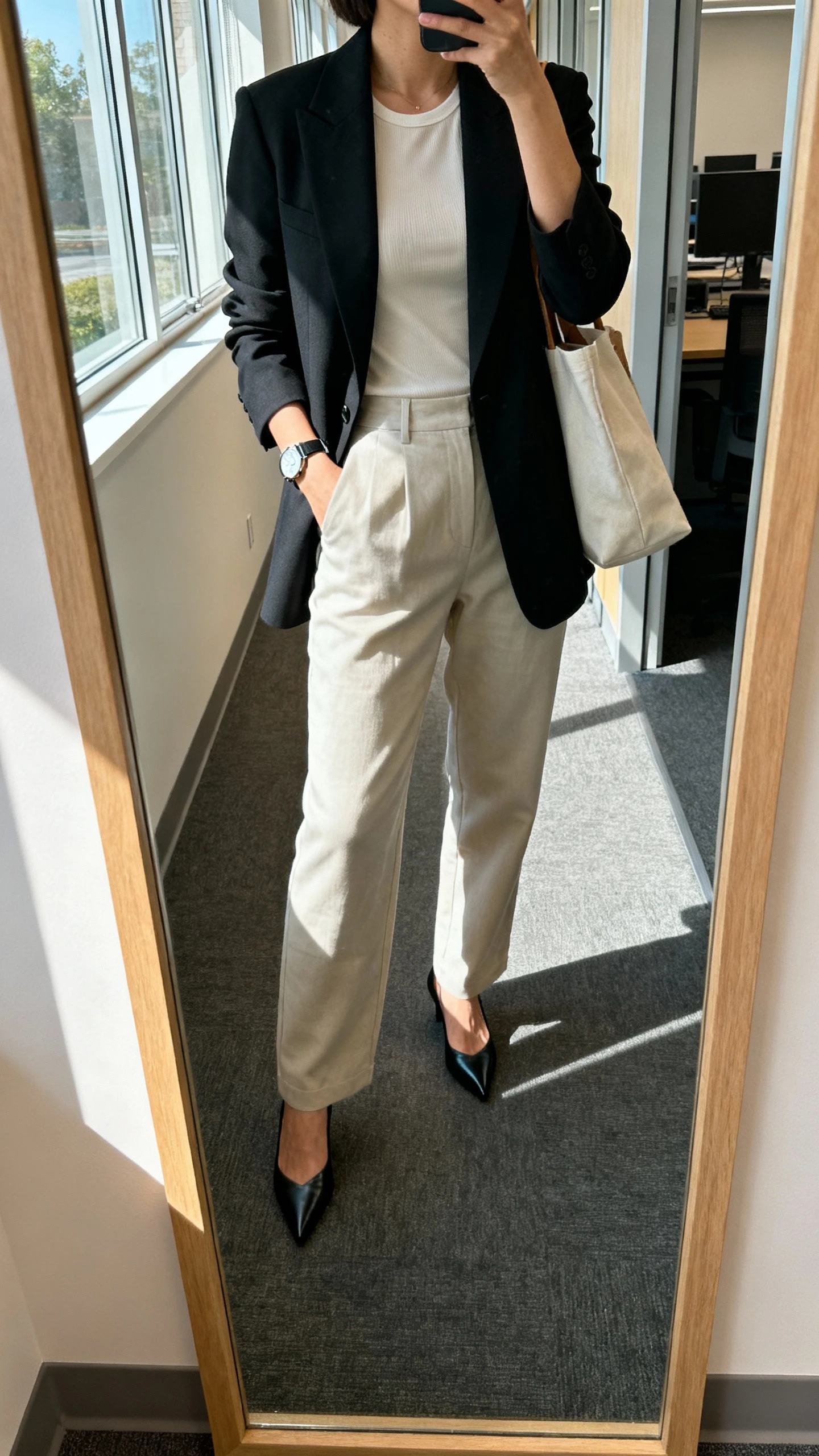 iPhone mirror selfie of a woman wearing a sharp blazer over a simple shell top with relaxed trousers and pointed heels, minimal watch and tote bag, face not visible, cropped at neck, office hallway mirror, morning window light, natural iPhone photo quality.