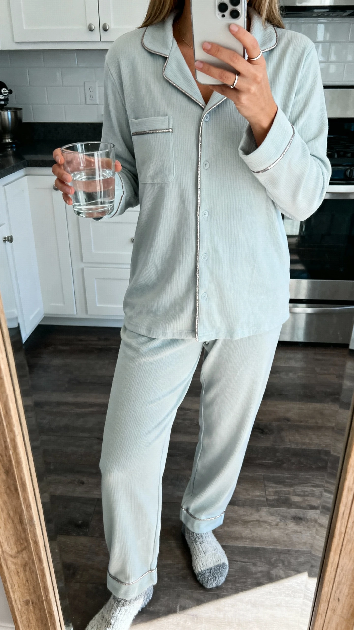 iPhone mirror selfie of a woman wearing an arctic-toned thermal pajama set with subtle metallic piping, cozy socks and a simple ring, face not visible, kitchen mirror reflection holding a glass of water, cool morning light, natural iPhone photo quality.