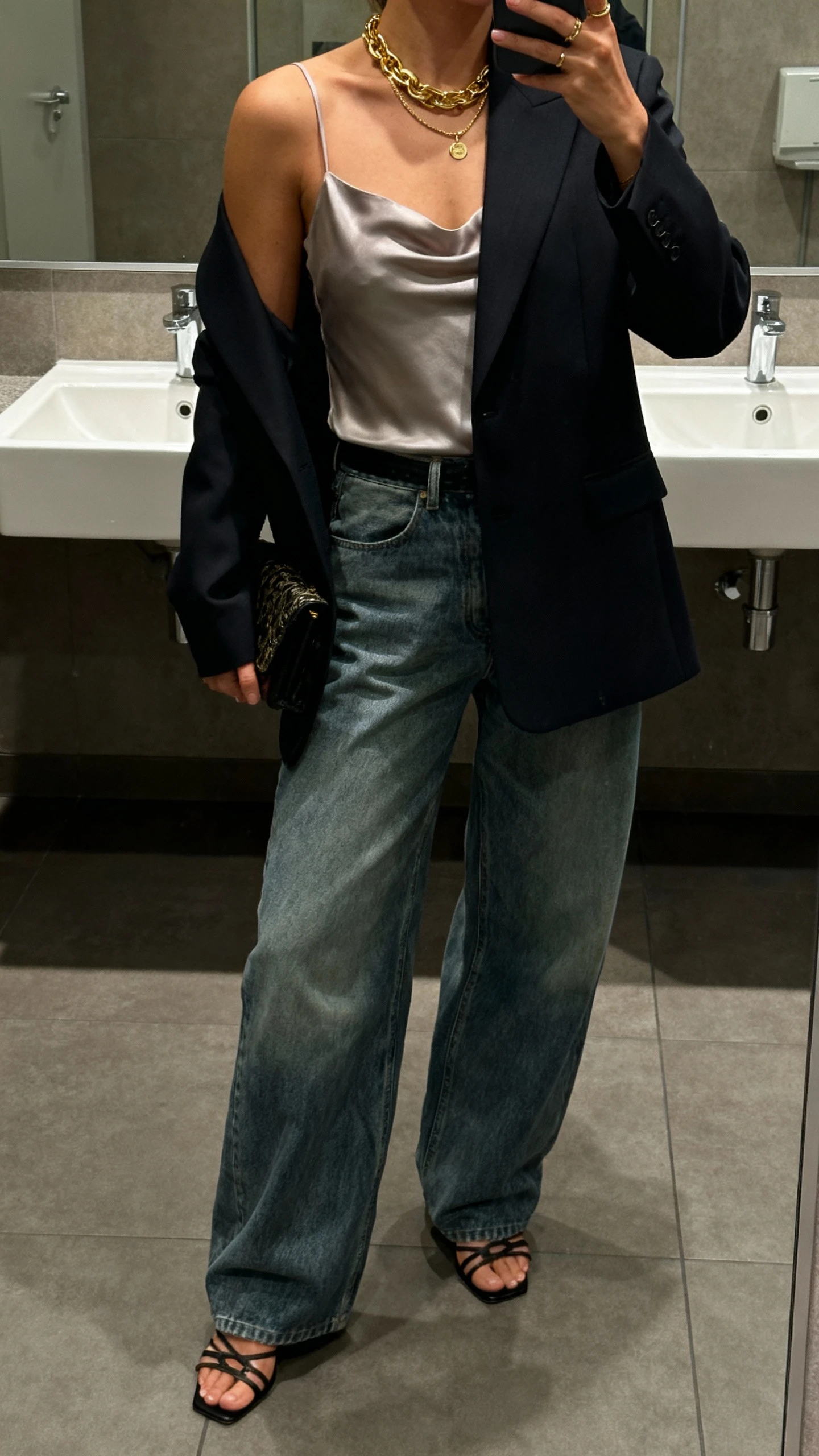 iPhone office-bathroom mirror selfie of a woman wearing baggy jeans, a sharp tailored blazer over a silky cami, strappy heels, layered gold jewelry and a small clutch, face not visible, cropped at shoulders, evening indoor lighting, natural iPhone photo