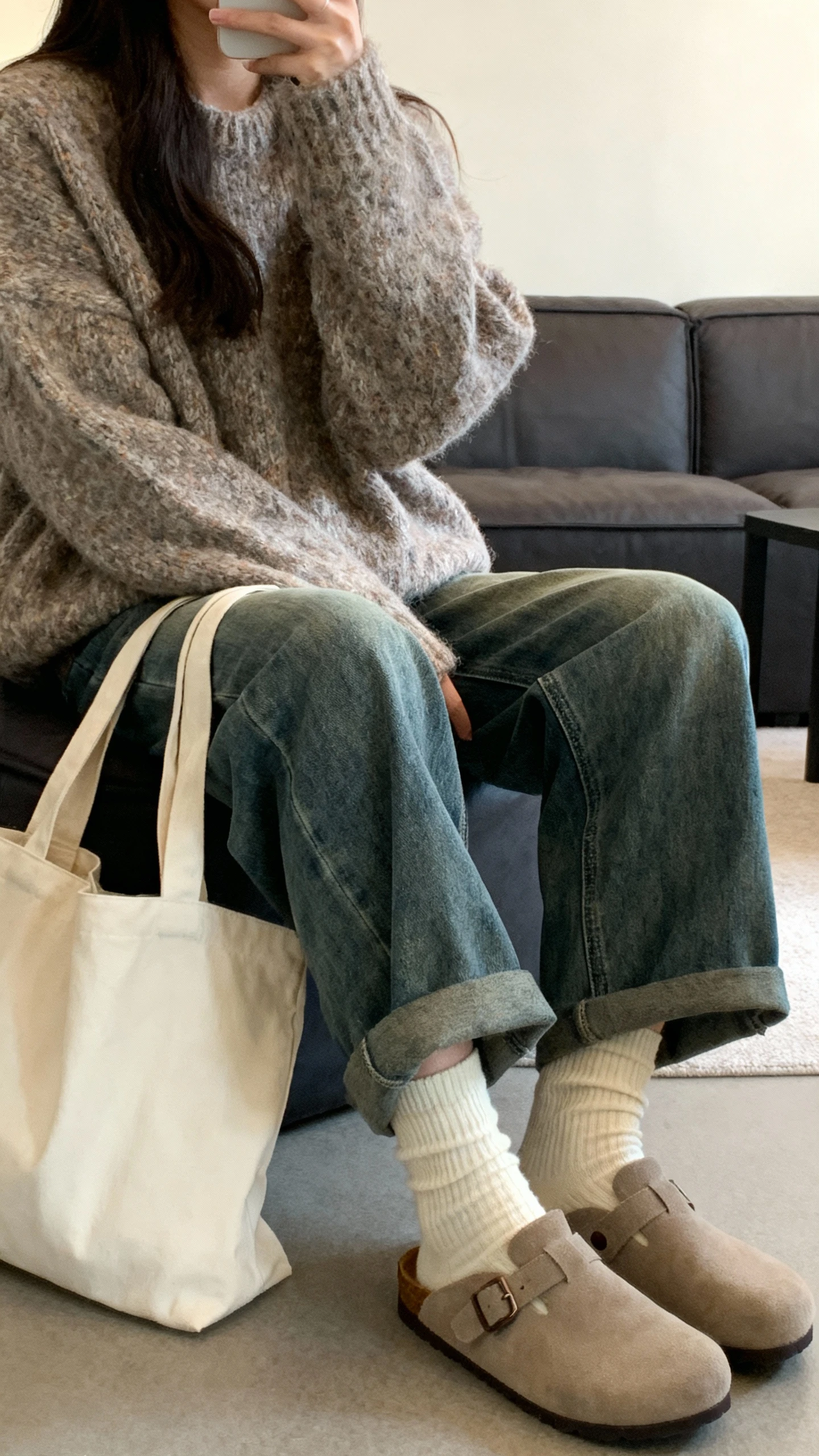 iPhone selfie from a side angle of a woman in baggy jeans, a cozy oversized chunky sweater, thick socks and slip-on clogs, simple tote bag, face not visible, living room couch setting with warm indoor light, casual iPhone aesthetic