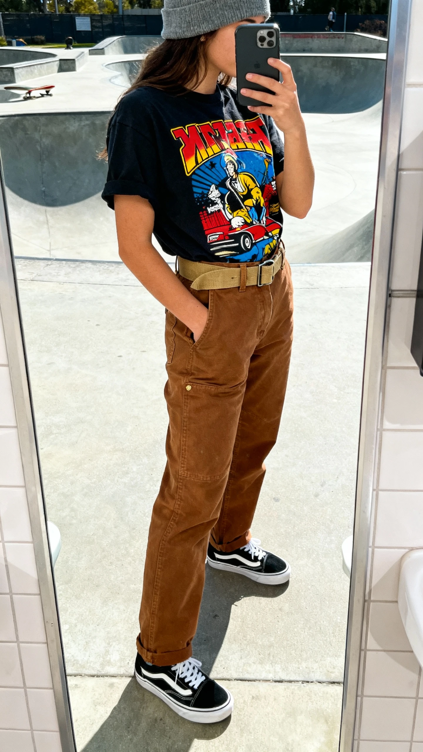 iPhone side-angle mirror selfie of a woman in a bold graphic tee tucked into brown workwear pants with a canvas belt, classic black-and-white Vans, beanie peeking in frame, face not visible, skate park restroom mirror, bright daylight, natural iPhone photo quality.