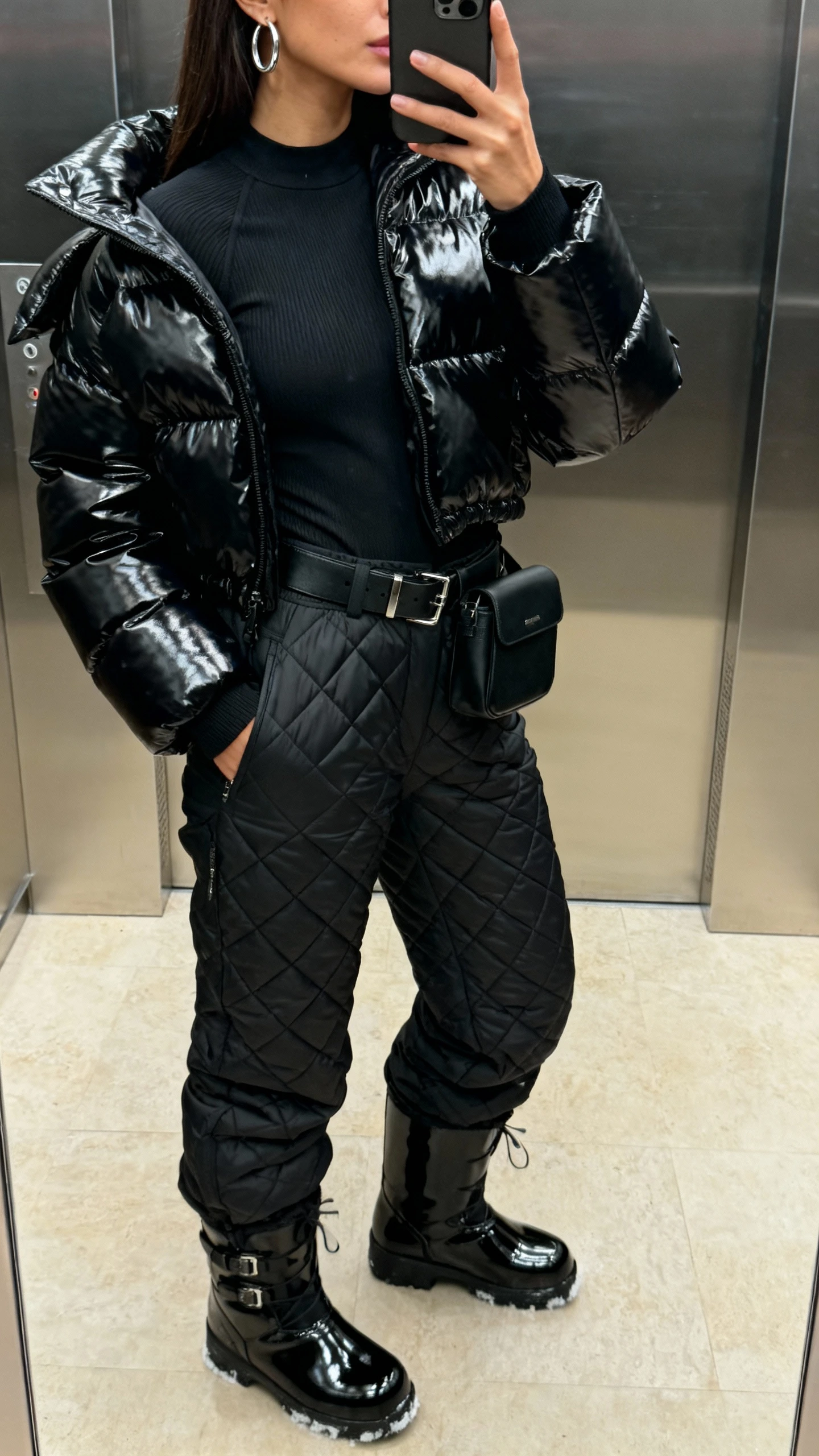 iPhone side-angle selfie of a woman in a chic blackout with mirror shine: glossy black puffer jacket, black thermal top, black quilted ski pants, patent leather snow boots, black belt bag, silver hoop earrings, face not visible, elevator mirror, soft indoor lighting, natural iPhone photo quality.