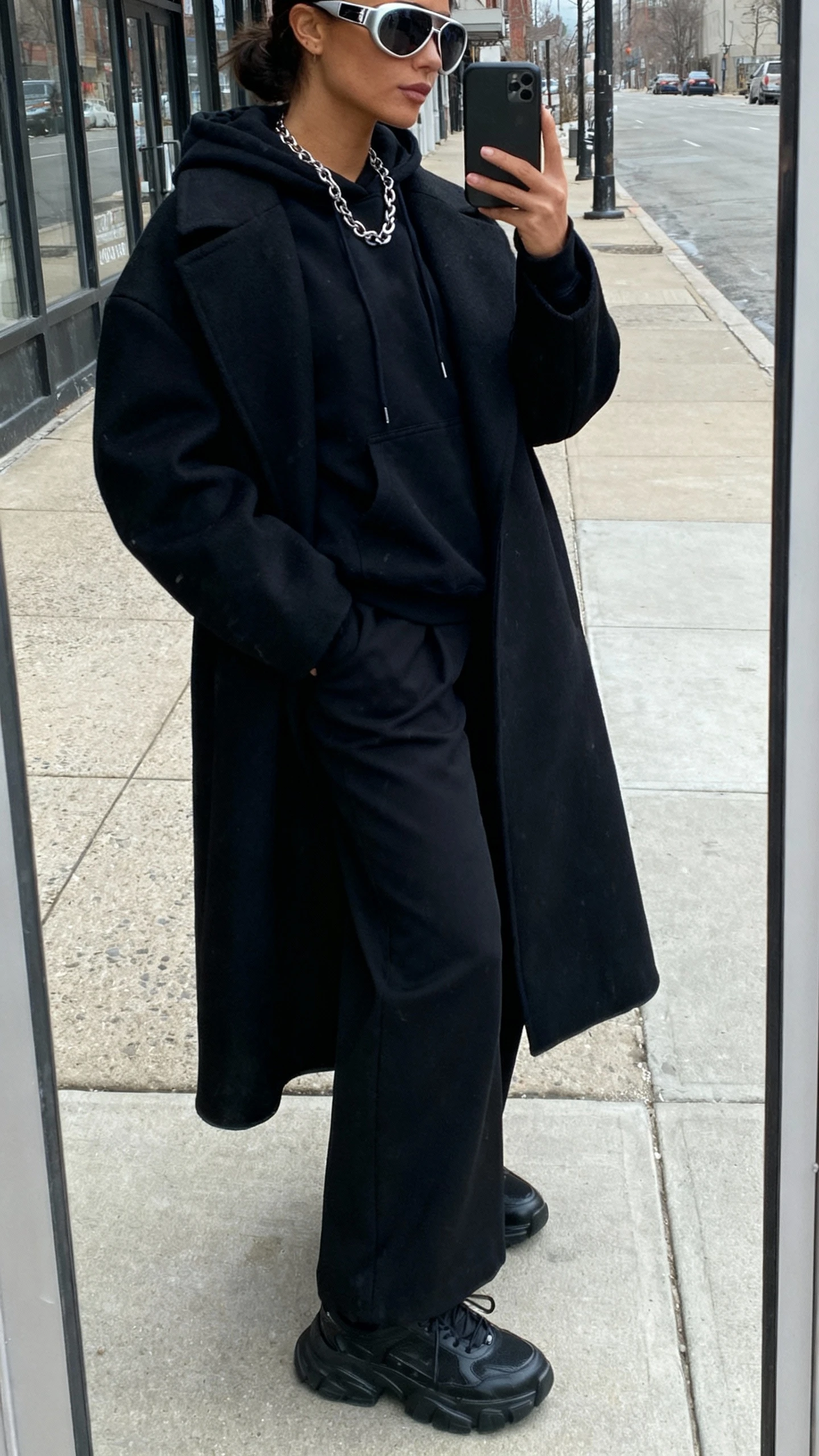 iPhone street mirror window selfie of a woman wearing oversized aviator sunglasses, monochrome black outfit: oversized black coat, black hoodie, black wide-leg trousers, black chunky sneakers, layered chain necklace, face not visible, side angle, overcast daylight on city sidewalk, casual iPhone quality