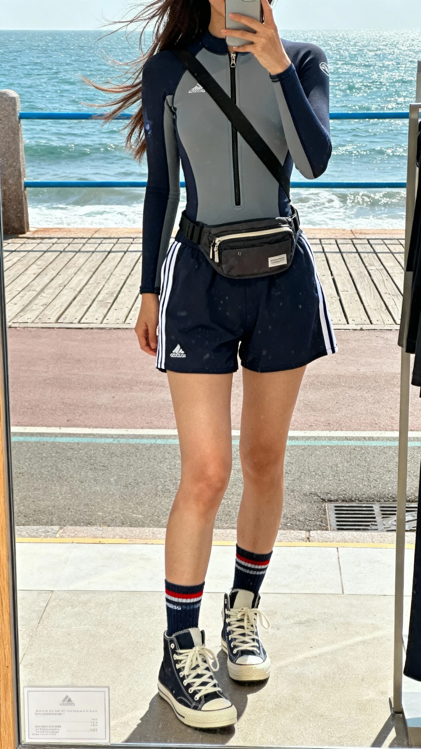 iPhone street-style mirror selfie (shop window) of a woman wearing a wetsuit-inspired zip top with track shorts, high-top sneakers, sporty socks, crossbody belt bag, face not visible, oceanfront boardwalk, breezy daylight, natural iPhone photo quality.