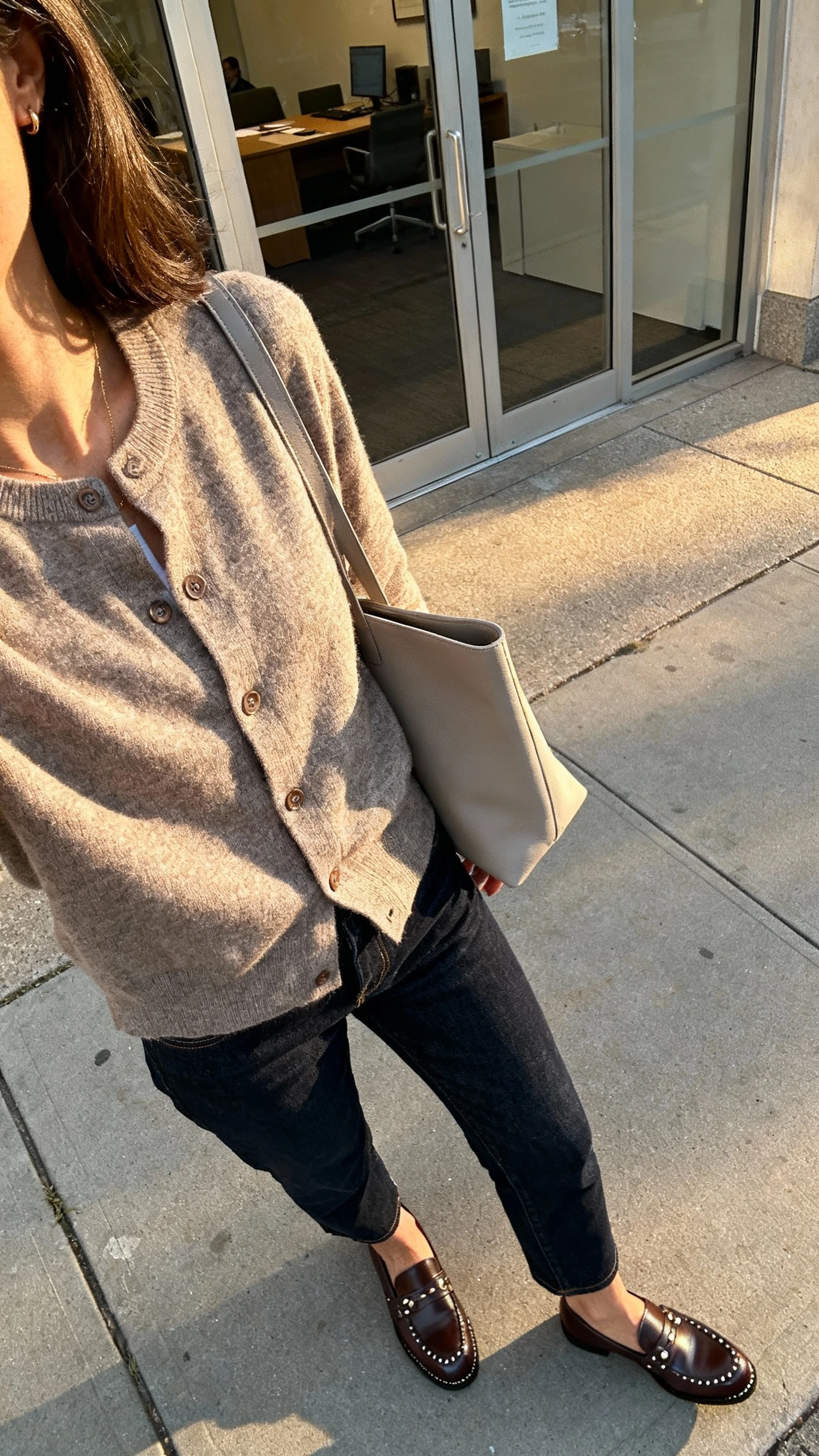 iPhone street-style selfie angled down from shoulder showing a woman wearing a cardigan jacket with dark denim, leather loafers, subtle studs and a minimalist tote, face not visible, sidewalk outside office, late afternoon natural light, casual iPhone shot.