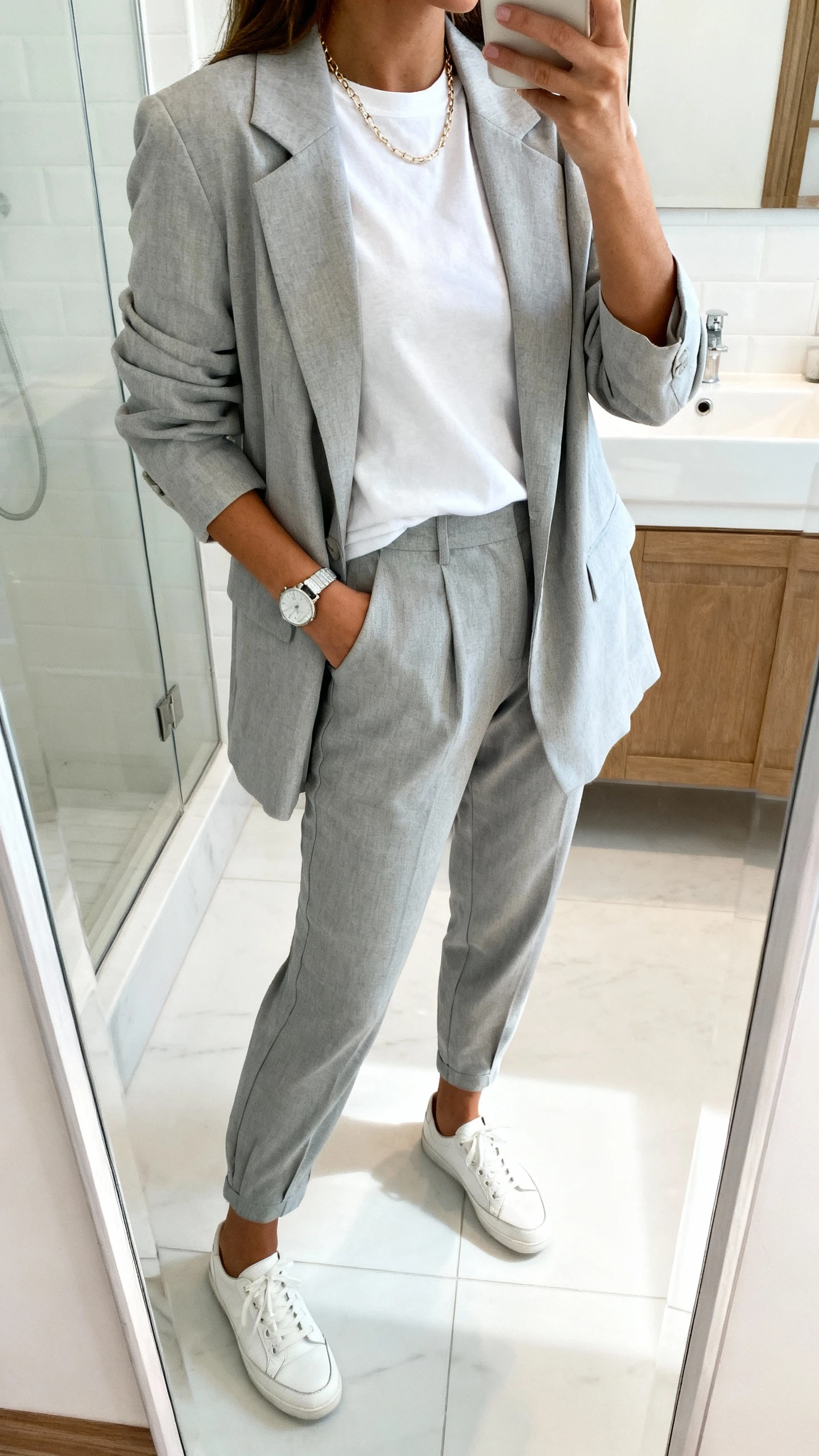 iPhone side-angle mirror selfie of a woman in a soft-power suit: light gray relaxed blazer with matching tapered trousers, white crewneck tee, clean white leather sneakers, delicate chain necklace, simple watch, face not visible, bathroom full-length mirror, bright indoor lighting, iPhone photo quality.