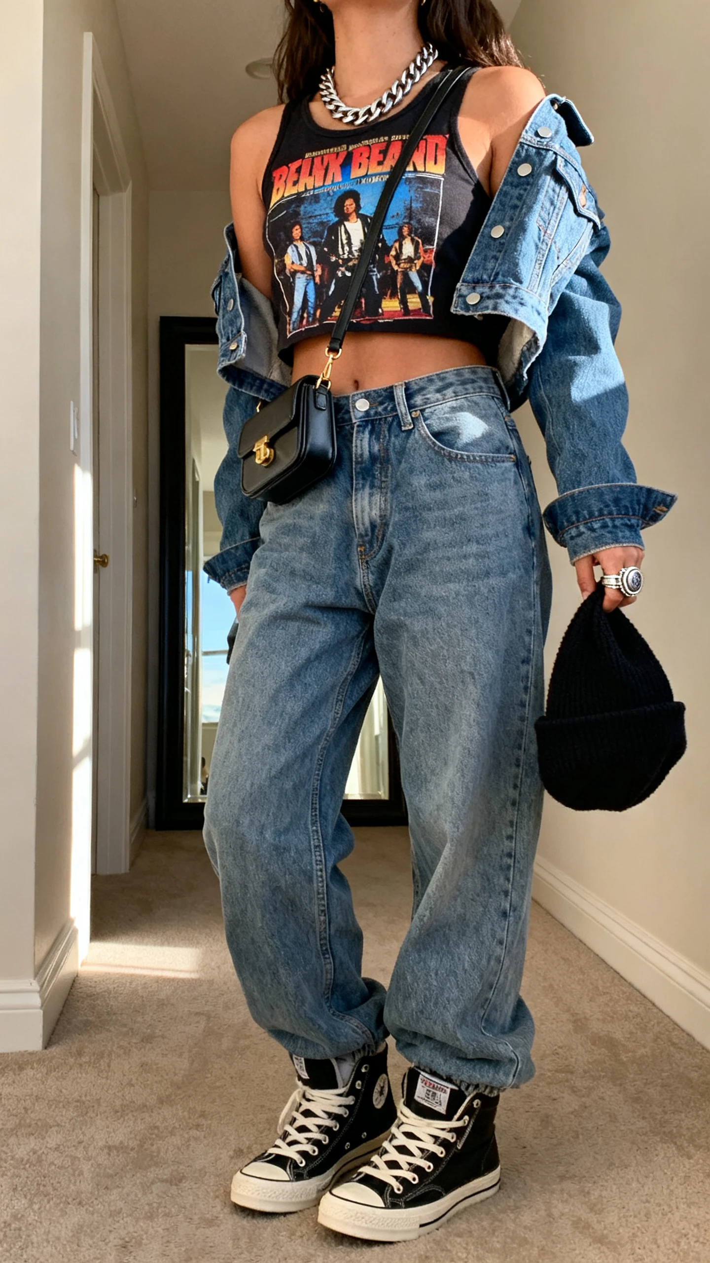 iPhone back-view candid of a woman wearing a graphic band tank, baggy dad jeans, a cropped denim jacket, high-top sneakers, a mini shoulder bag, a chunky chain necklace, and a statement ring, with a beanie in hand; face not visible, hallway mirror, natural afternoon light, iPhone photo quality.