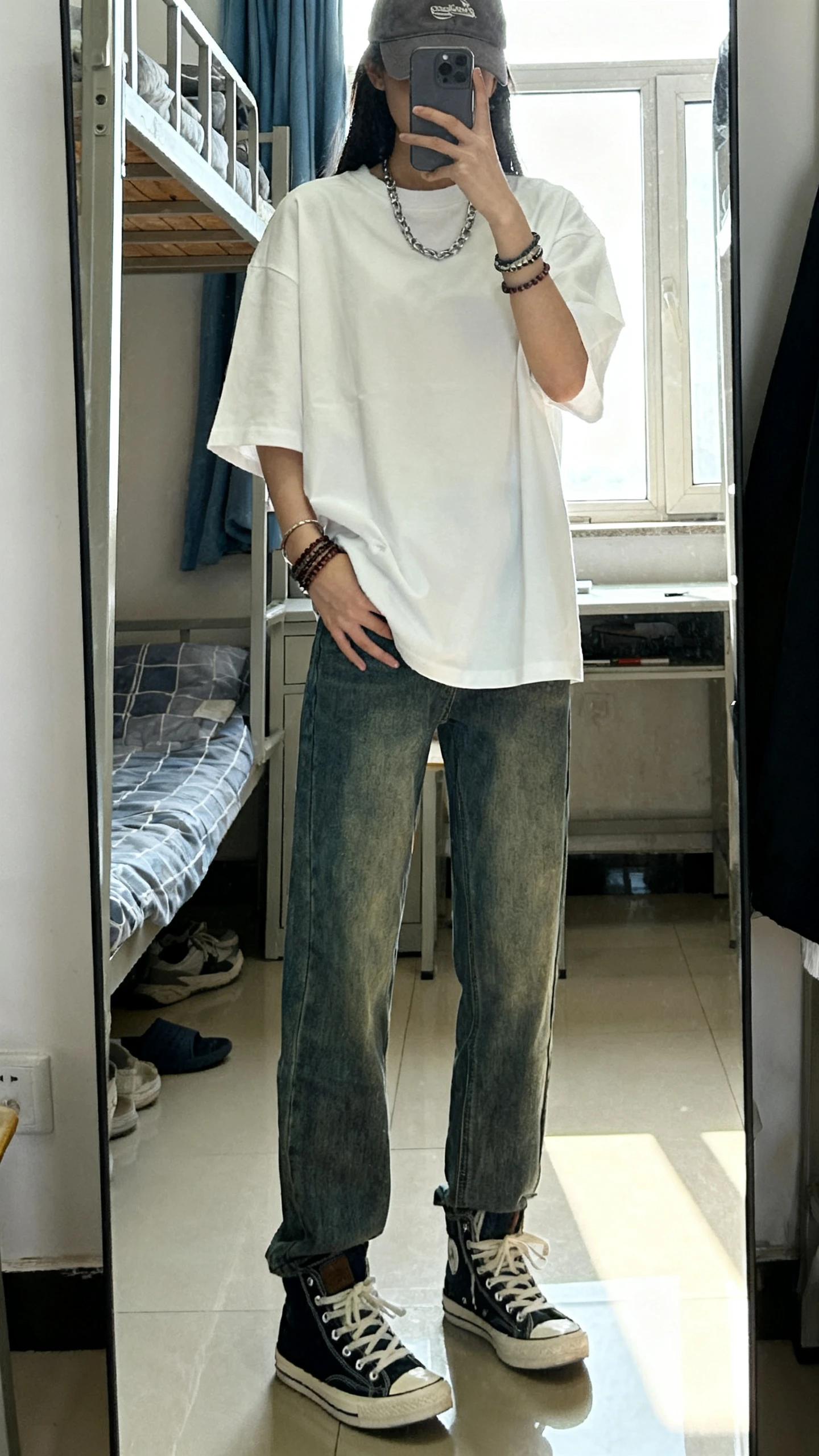 iPhone mirror selfie of a woman in a boxy white tee tucked into straight-leg medium-wash jeans, high-top sneakers, a baseball cap, a simple chain necklace, and stacked bracelets, face not visible, cropped at shoulders, dorm bedroom mirror, morning window light, iPhone photo quality.