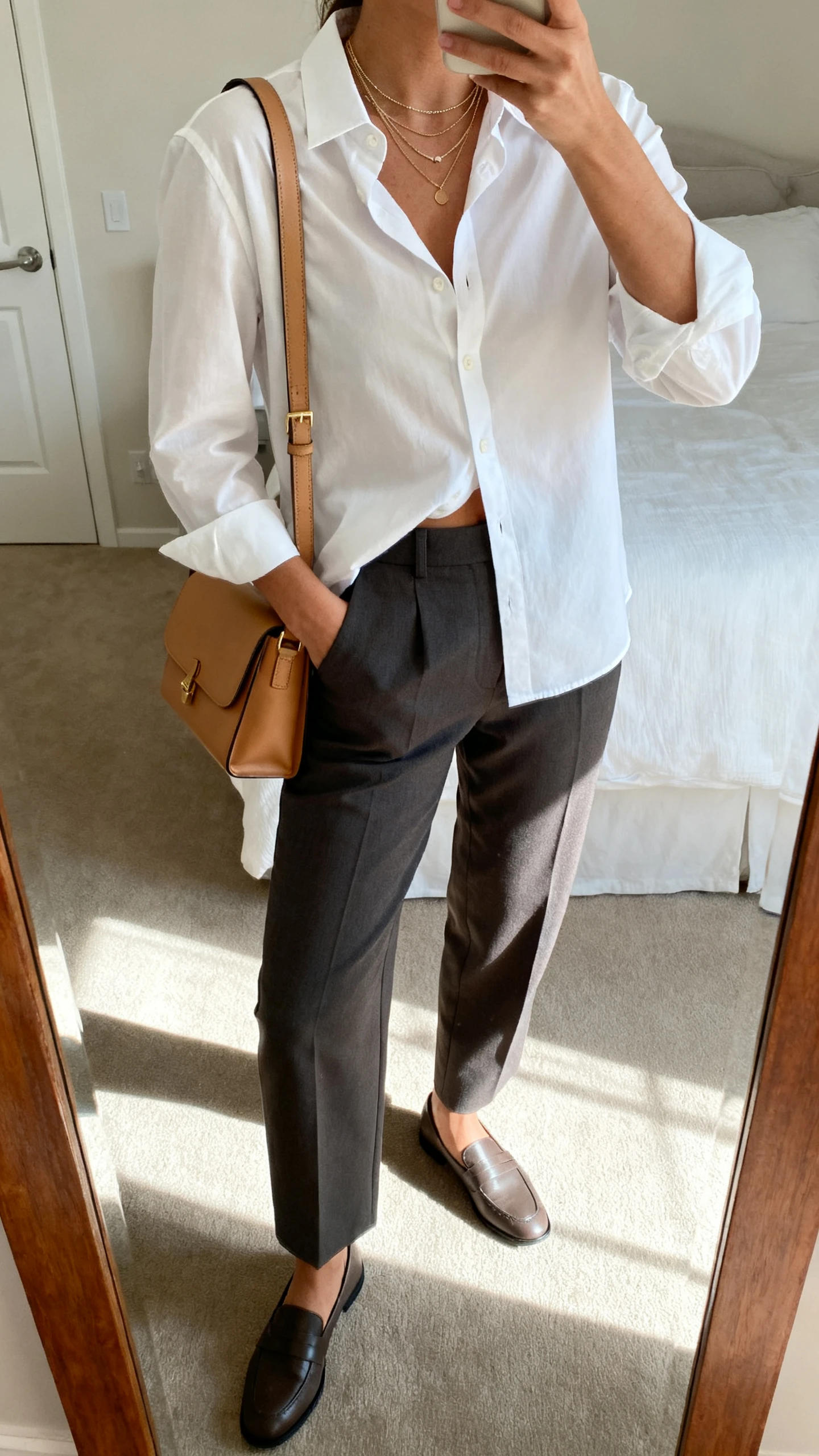 iPhone mirror selfie of a woman in a crisp white button-down shirt, half-tucked into tailored charcoal trousers, with a tan leather shoulder bag, loafers, and delicate layered necklaces, face not visible, cropped at shoulders, bedroom mirror, natural daylight, iPhone photo quality