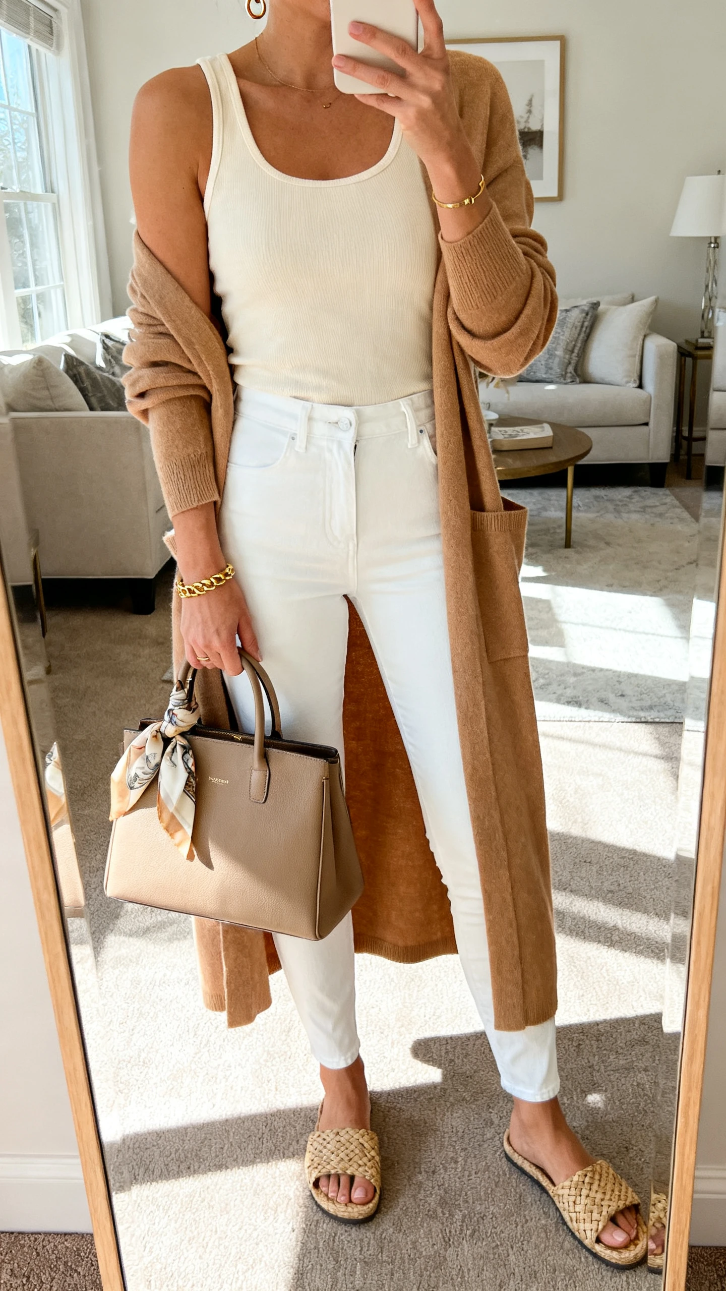 iPhone mirror selfie of a woman in a neutral cream tank, thick white slim jeans, a camel longline cardigan-coat, woven slides, a structured tote with a silk scarf on the handle, gold bracelets and small hoops; face not visible, cropped at neck, living-room full-length mirror, bright daylight, iPhone photo quality.