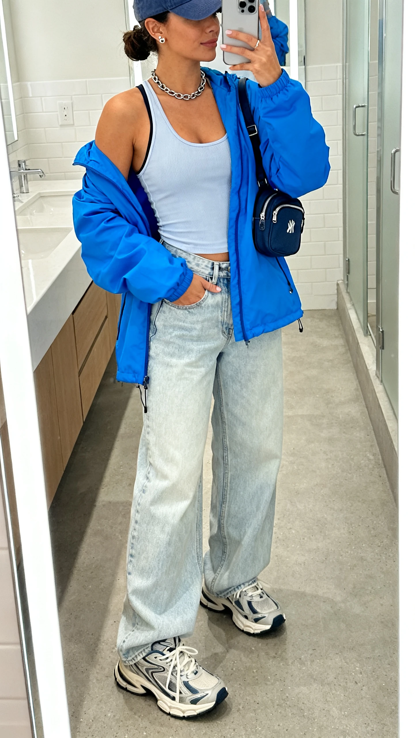iPhone mirror selfie of a woman in a racerback performance tank, light-wash wide-leg jeans, a punchy-color windbreaker (electric blue), chunky retro trainers, a baseball cap, a mini backpack, minimal stud earrings, and a chain necklace; face not visible, bathroom mirror, bright indoor lighting, iPhone photo quality.