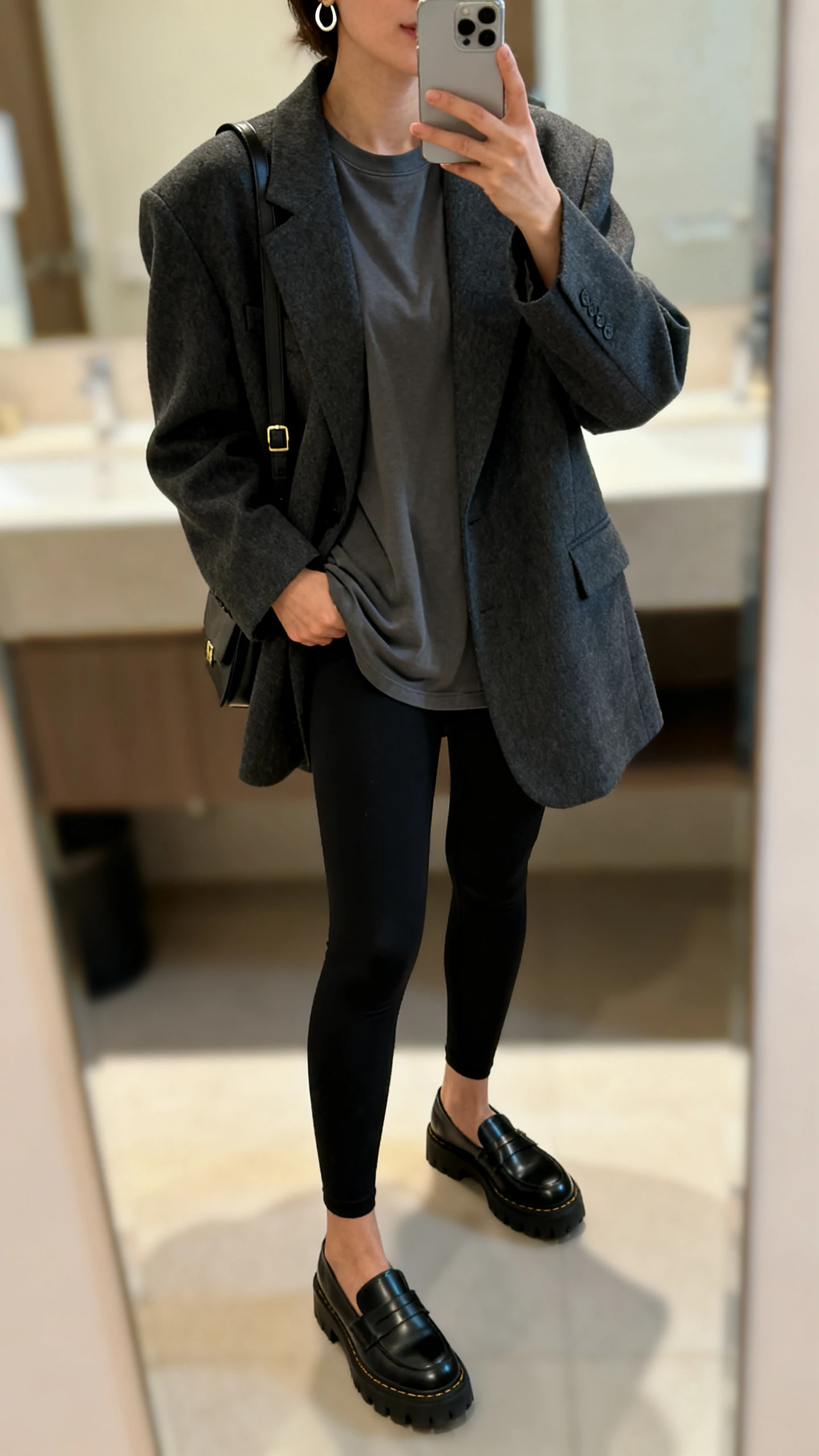 iPhone mirror selfie of a woman in an oversized charcoal blazer over a basic crewneck tee, thick black leggings, chunky loafers, a shoulder bag, and small hoop earrings, face not visible, bathroom mirror, indoor lighting, iPhone photo quality.