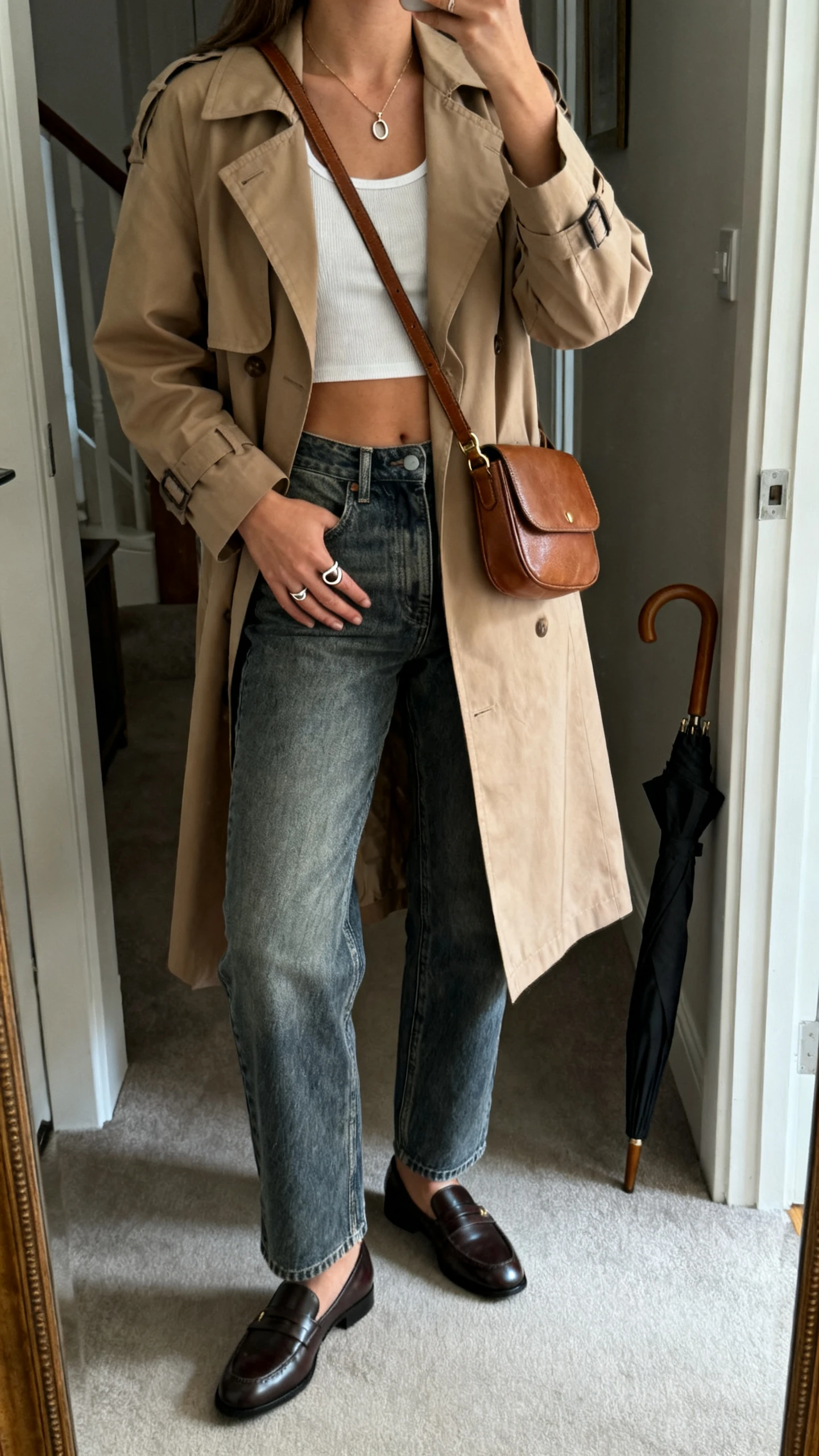 iPhone mirror selfie of a woman wearing a cropped tank, high-rise mid-wash straight jeans, a light tan trench coat, sleek loafers, a leather crossbody, a simple pendant necklace, a signet ring, and an umbrella leaning nearby; face not visible, cropped at neck, entryway mirror, overcast indoor light, iPhone photo quality.