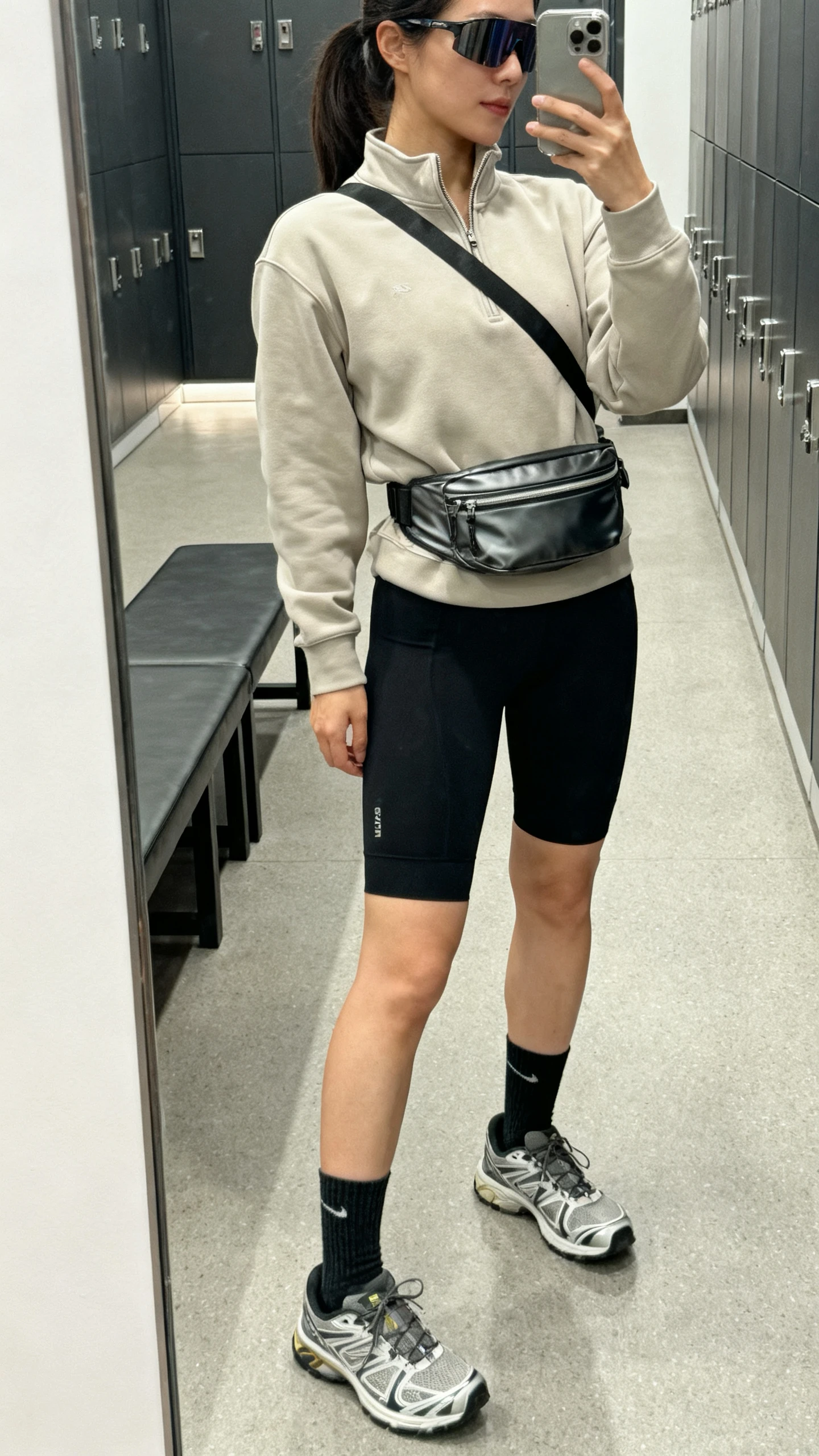 iPhone side-angle mirror selfie of a woman wearing a neutral quarter-zip sweatshirt, black bike shorts, running sneakers, crew socks, a sleek crossbody belt bag, and sporty sunglasses, face not visible, gym locker room mirror, indoor lighting, iPhone photo quality.