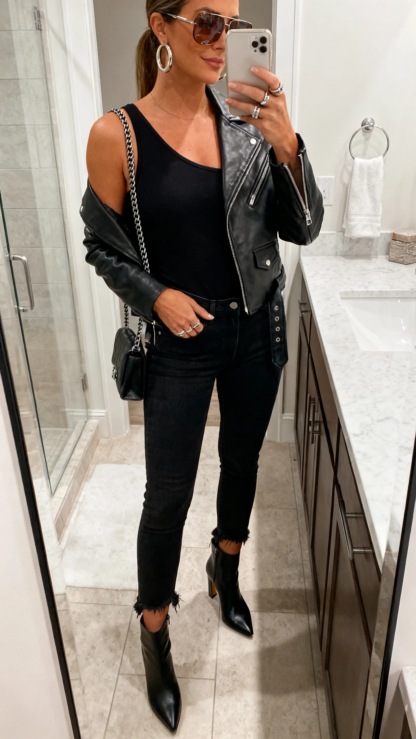 Side-angle iPhone mirror selfie of a woman wearing an asymmetrical black tank, black slim jeans with raw hem, a leather moto jacket, heeled ankle boots, a chain-strap shoulder bag, chunky hoops, stacked rings, and smoky sunglasses; face not visible, bathroom mirror, indoor lighting, iPhone photo quality.